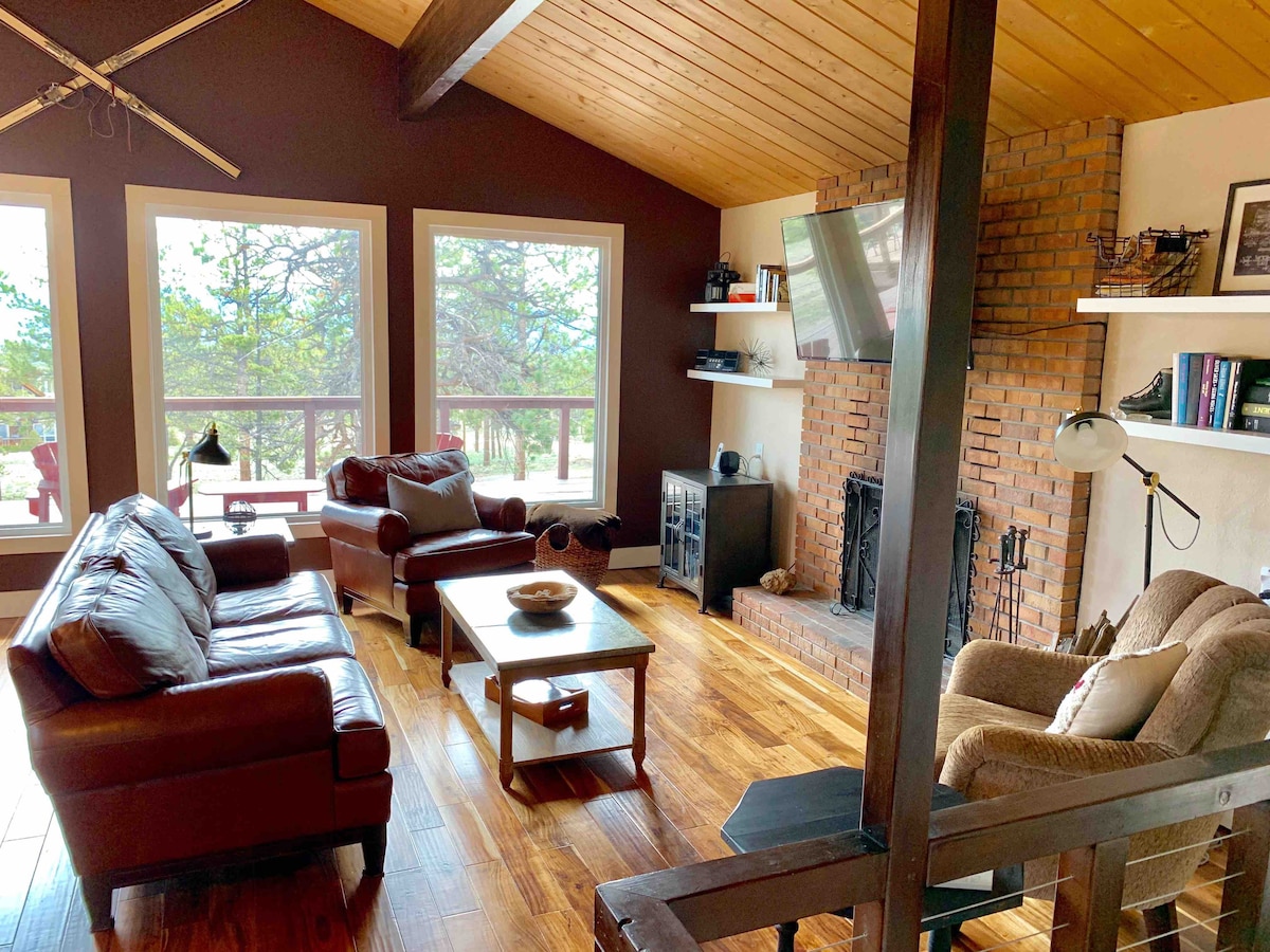 A spacious living room is furnished with a leather sofa, two armchairs, and a wooden coffee table. Large windows provide natural light and views of the surrounding woods. A brick fireplace serves as a focal point, complemented by a mounted television and bookshelves.