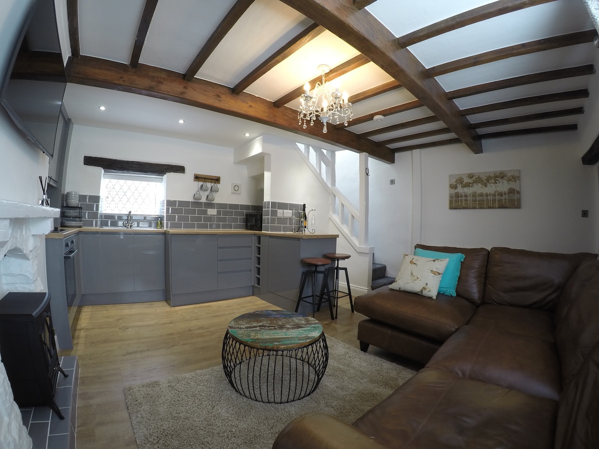 The open-plan living area showcases a cozy leather corner sofa and a round coffee table on a soft rug. Exposed wooden beams enhance the rustic charm. The kitchen features contemporary cabinetry and modern appliances, with warm lighting creating an inviting atmosphere.