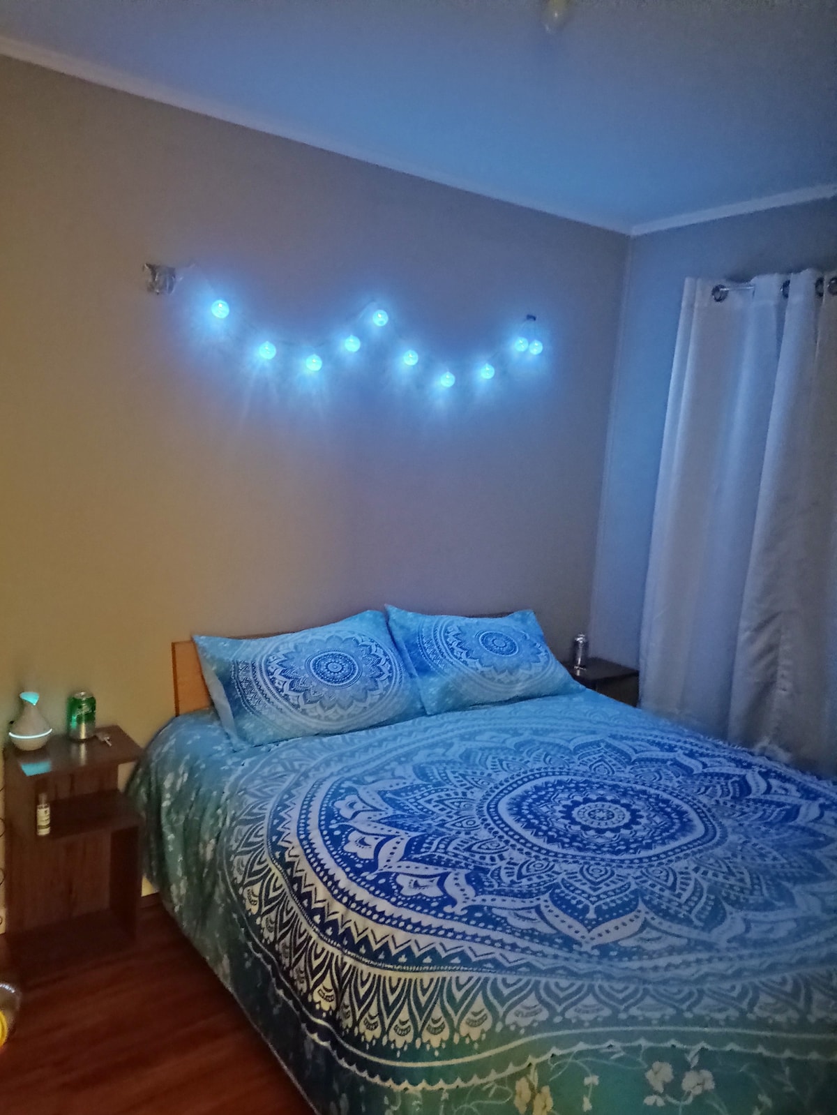 A cozy bedroom features a spacious bed adorned with a blue and white patterned comforter. Soft blue lights are strung above the bed, creating a calming ambiance. Nightstands are placed on either side, complementing the serene decor. Natural light filters through the white curtains.