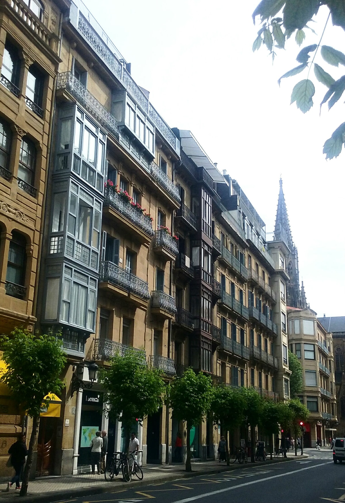 A street view showcases a classic building facade featuring intricate detailing and balconies adorned with plants. The image captures a blend of architectural styles, with sunlight highlighting the warm tones of the exterior. A church spire is visible in the background, adding to the historic atmosphere.