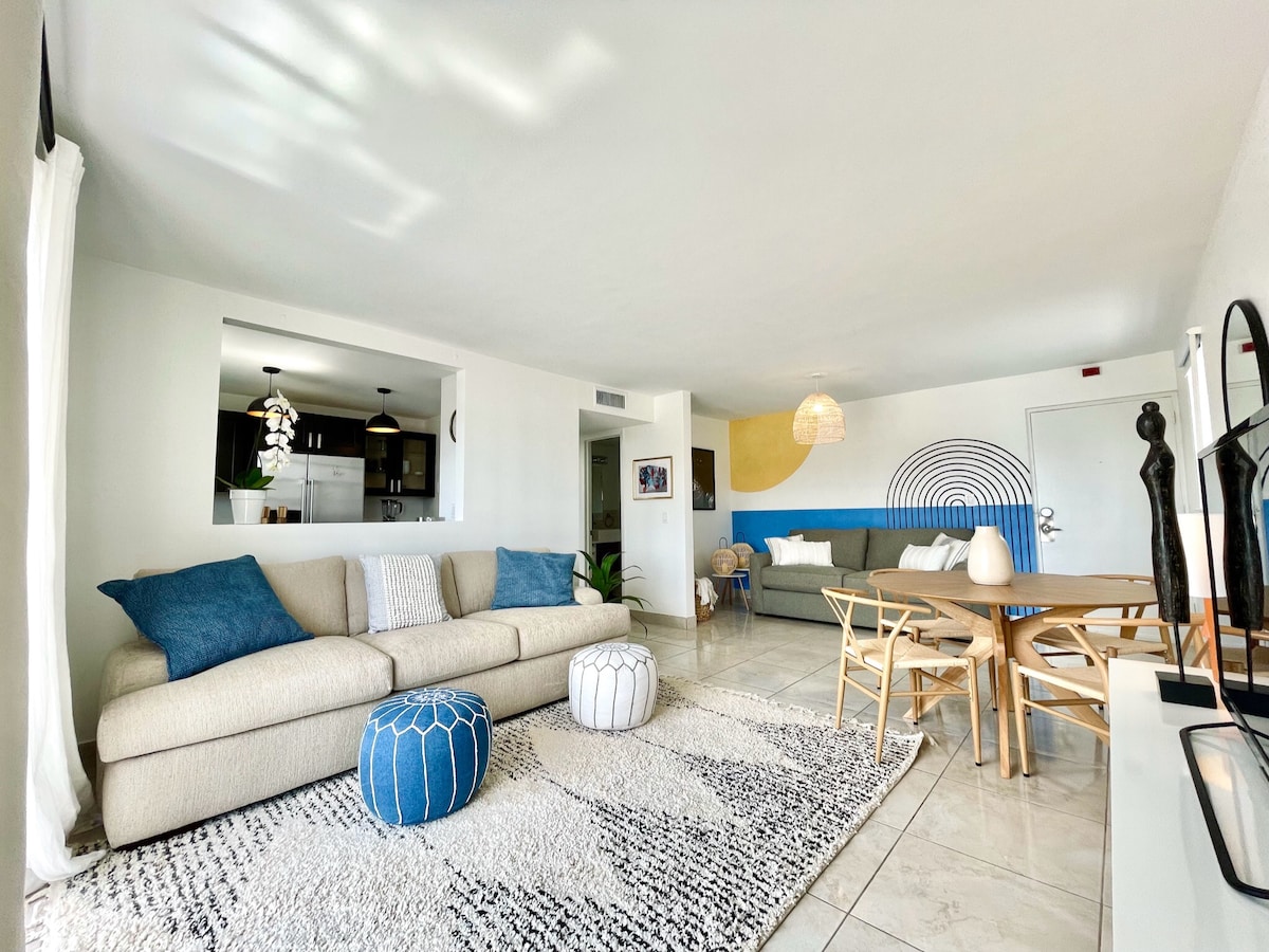 A bright and open living area features a spacious light-colored sofa complemented by blue accent pillows. A round coffee table sits on a textured rug, while a dining table with wooden chairs occupies the space. A kitchen area is partially visible in the background.