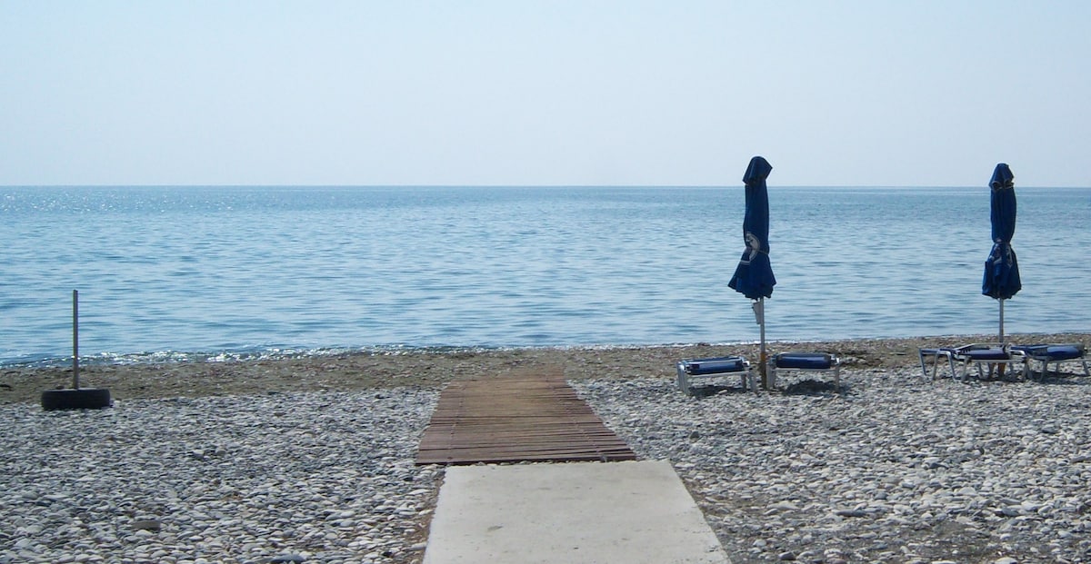 A wooden pathway leads from a pebbled beach to calm waters. Two blue umbrellas are positioned over lounge chairs, providing shade. The horizon displays a serene sky meeting the gentle sea, creating a tranquil coastal scene.
