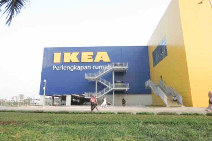 The exterior of an IKEA store is displayed, characterized by its large, colorful facade featuring blue and yellow elements. A wide staircase leads to the entrance, and people can be seen walking in the vicinity. Lush green grass frames the lower portion of the image.