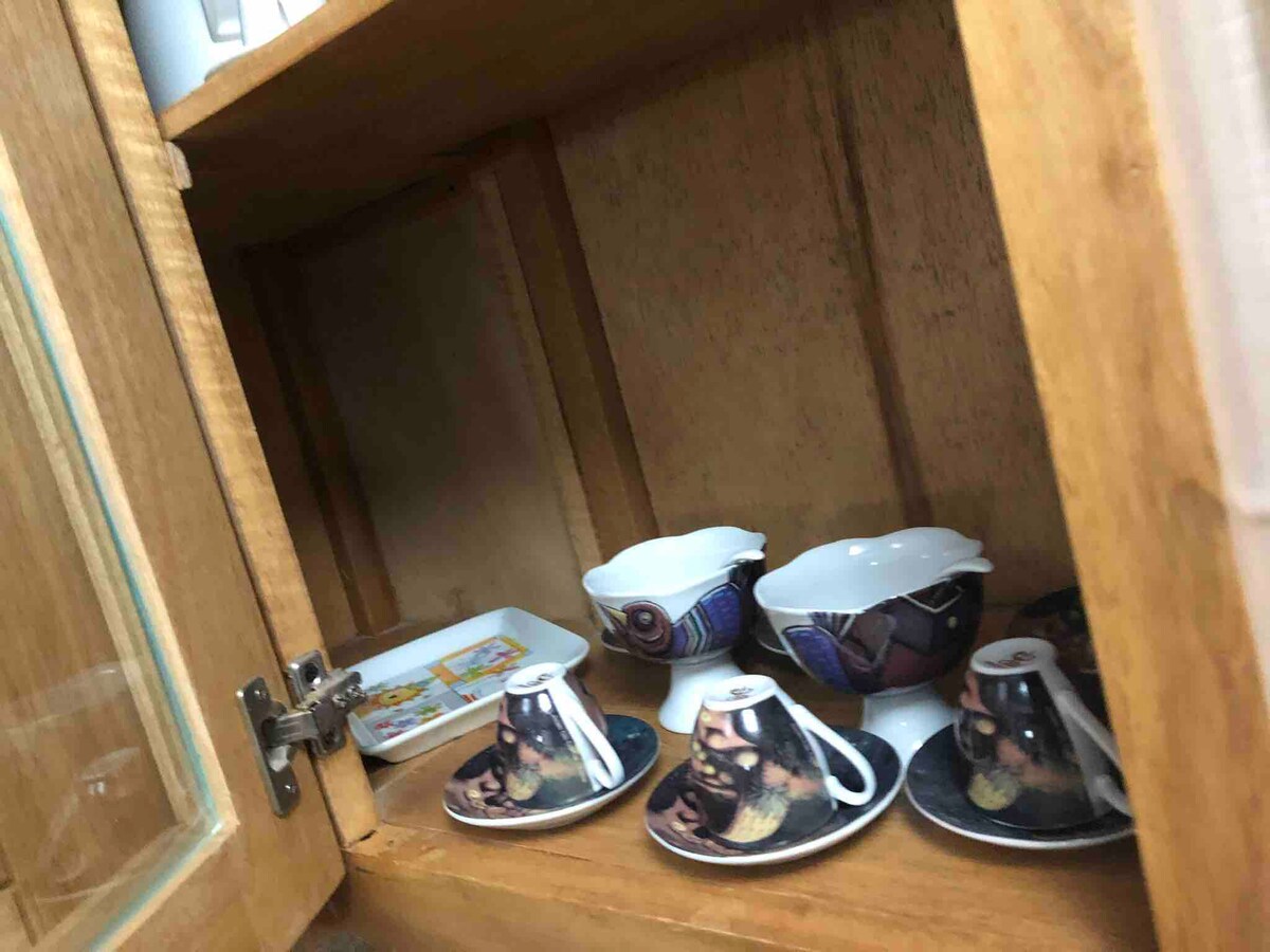 A wooden cupboard contains various ceramic items, including small coffee cups with matching saucers and a creamer set. The interior is illuminated, highlighting the colorful patterns on the dishes. A small tray is positioned at the back of the cupboard.