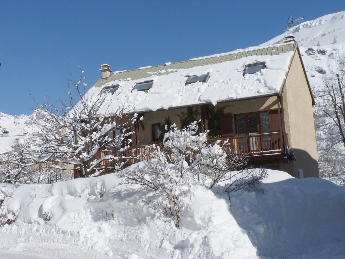 A chalet-style building is covered in fresh snow, with a sloped roof and skylights evident. Surrounding trees are blanketed in snow, and a wooden balcony extends from the front. The clear blue sky above contrasts with the winter scenery.