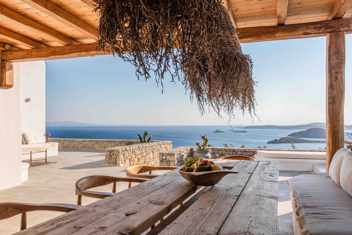 An outdoor dining area features a large wooden table surrounded by curved seating. The space is shaded by a natural wood pergola with hanging foliage, offering a view of the sea and distant islands. Coastal plants are arranged along the terrace, creating a serene environment.