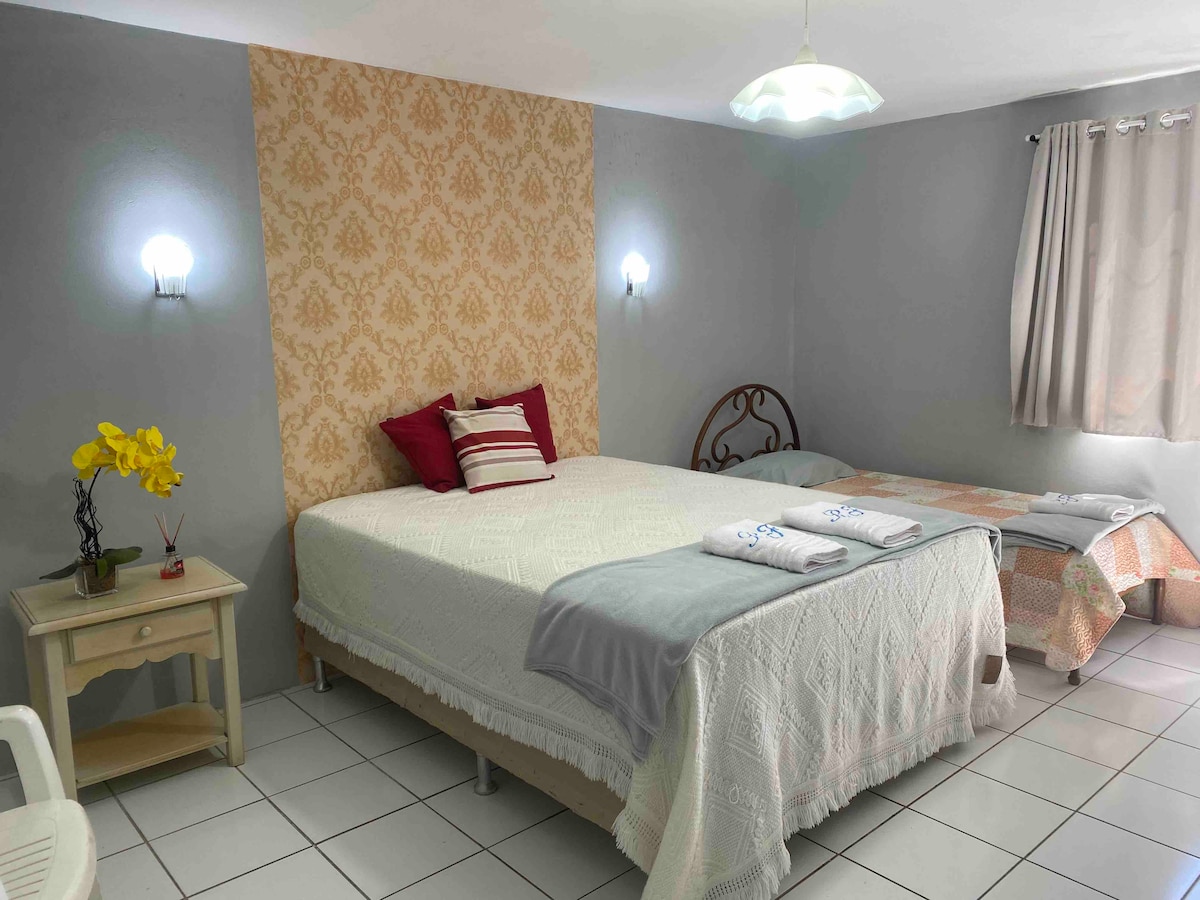 A cozy sleeping area features a spacious bed adorned with a light-colored quilt and decorative pillows. A single bed is positioned nearby, with towels neatly arranged. Soft lighting emanates from wall sconces, while a small table holds a vibrant yellow flower arrangement.