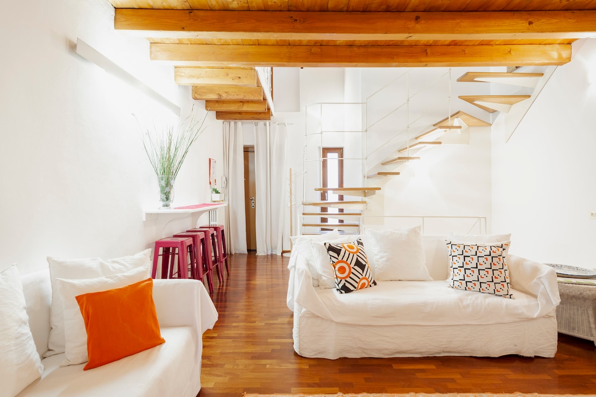 A bright living area features white sofas with colorful accent pillows, complemented by warm wooden beams overhead. A stylish staircase leads to an upper level. The space includes a sleek bar area with high stools, enhancing the open feel of the room.