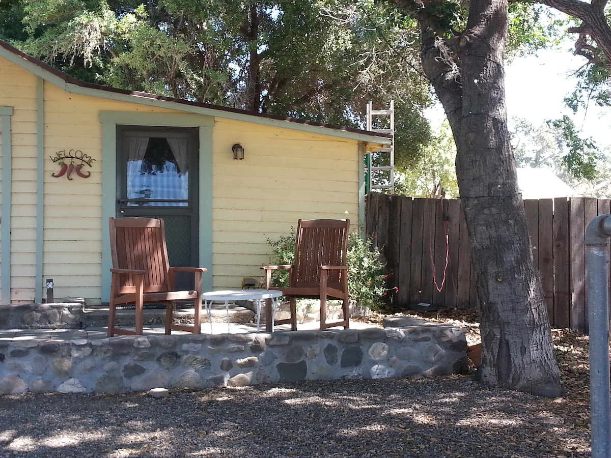 A cozy front patio is featured with two wooden chairs and a small table, set against a backdrop of a rustic cottage. The area is shaded by a large tree, surrounded by gravel and a stone border.
