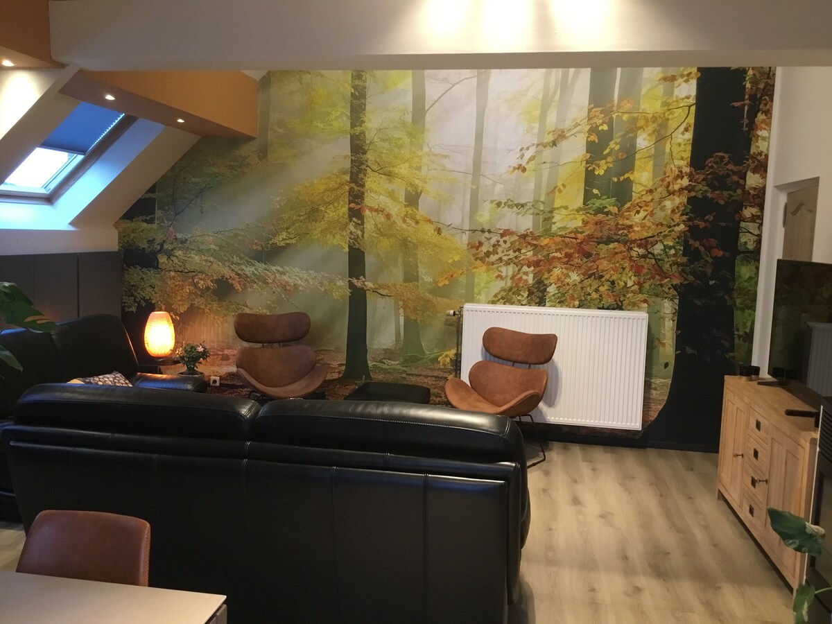 A cozy living area features a large black sofa and two brown armchairs, positioned in front of a wooden sideboard. A forest mural with soft light filtering through trees creates a serene backdrop. A radiator is also visible, adding functionality to the space.