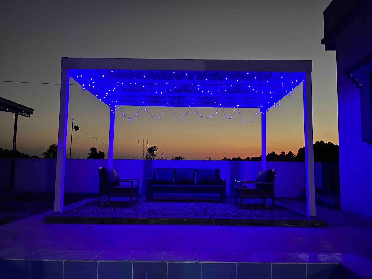 An outdoor seating area is illuminated by blue string lights against a twilight backdrop. Two chairs and a sofa are arranged under a white pergola, creating a cozy space. A quiet landscape is visible in the distance, adding to the serene atmosphere.