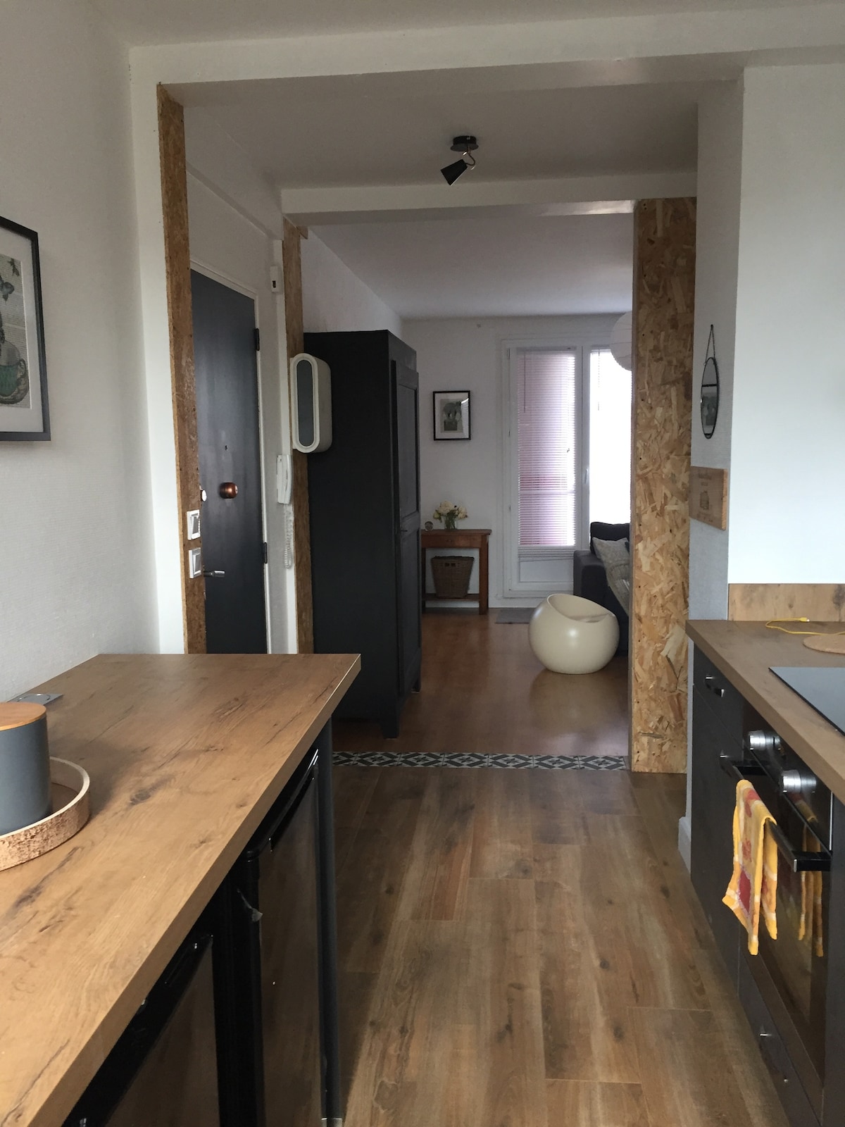 The image captures a view of a modern kitchen featuring wooden countertops and sleek cabinetry. Soft natural light enters from a nearby window, illuminating the space. In the background, a hallway leads to a cozy living area, framed by stylish decorations and wooden accents.