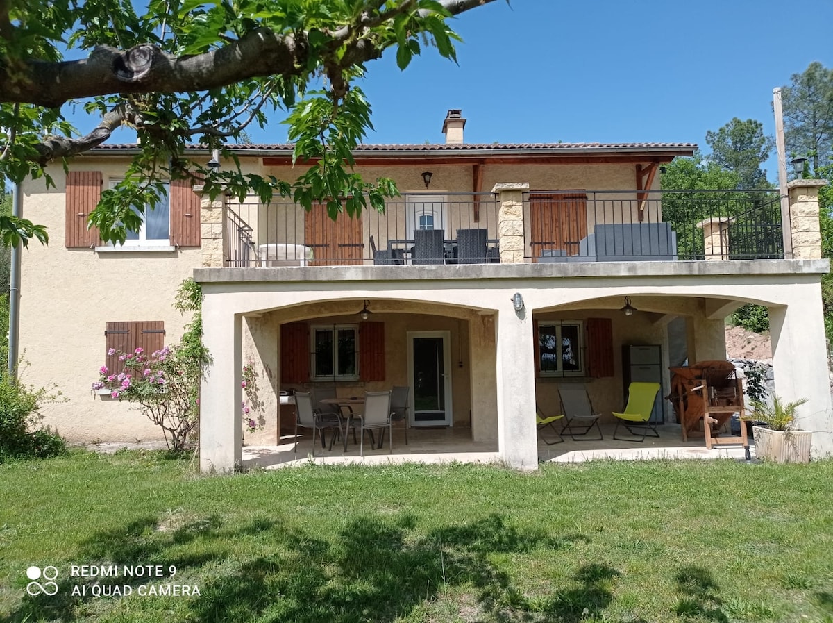 The villa's exterior features a combination of warm-toned walls and classic wooden shutters. A covered patio is visible, adorned with outdoor furniture for relaxation. The upper balcony provides additional seating, while greenery frames the peaceful setting.