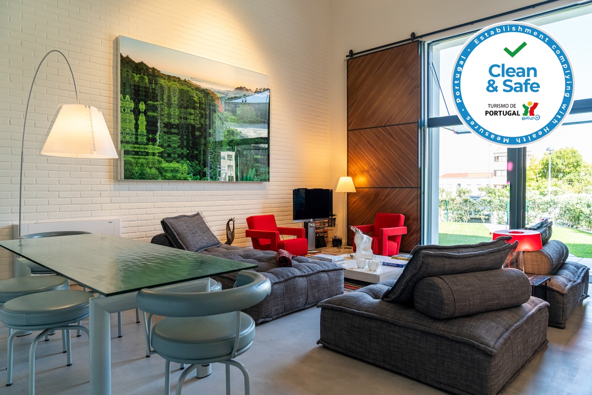 A spacious living area features contemporary furniture with cozy seating, including a pair of red accent chairs and comfortable grey sofas. Large windows allow natural light to fill the room, with a view of the garden outside. A dining table is positioned near the seating area.