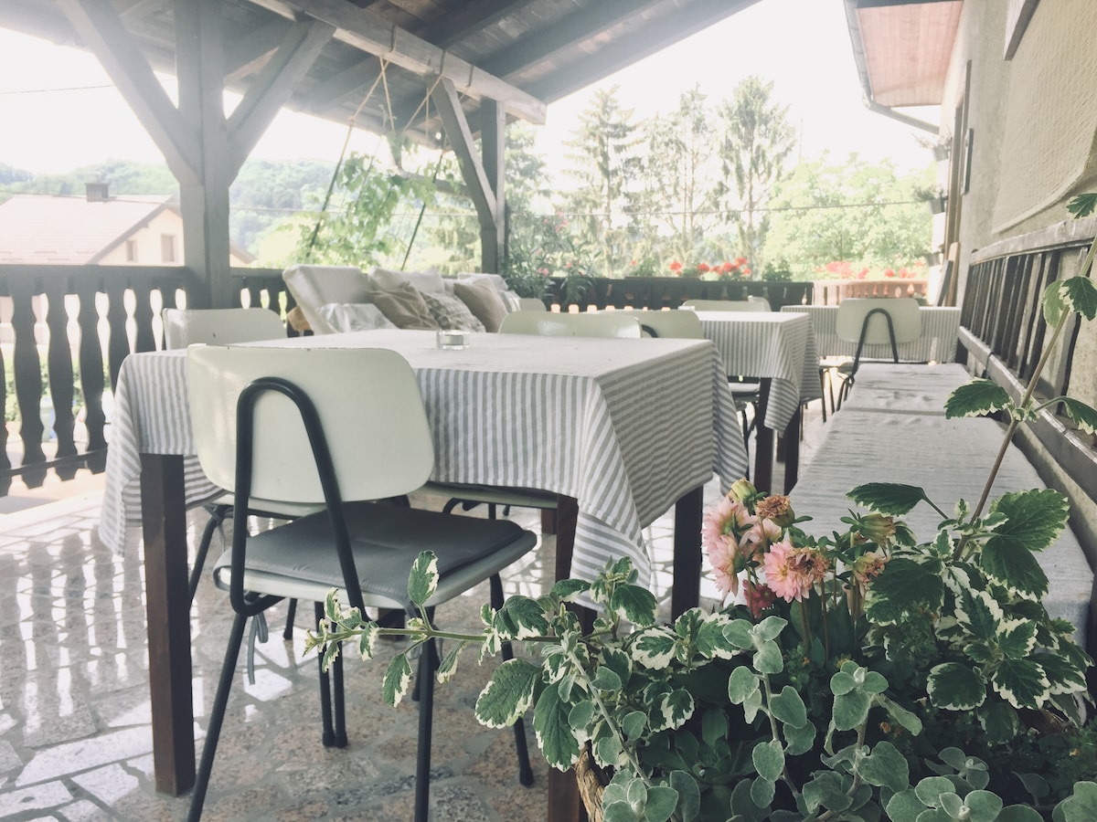 A spacious terrace features several tables covered with striped tablecloths. Comfortable chairs are arranged around the tables, while planters with greenery and blooming flowers add natural beauty. The area is shaded by a wooden roof, creating a relaxed outdoor setting.