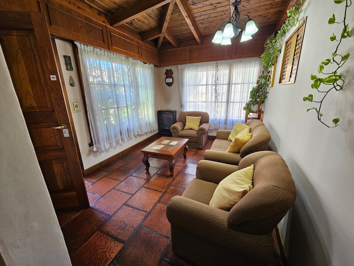 The living area features warm wood beams overhead and a combination of natural light from large windows with sheer curtains. Two comfortable armchairs are positioned around a wooden coffee table, inviting relaxation. Green plants are placed in window ledges, adding a touch of nature.