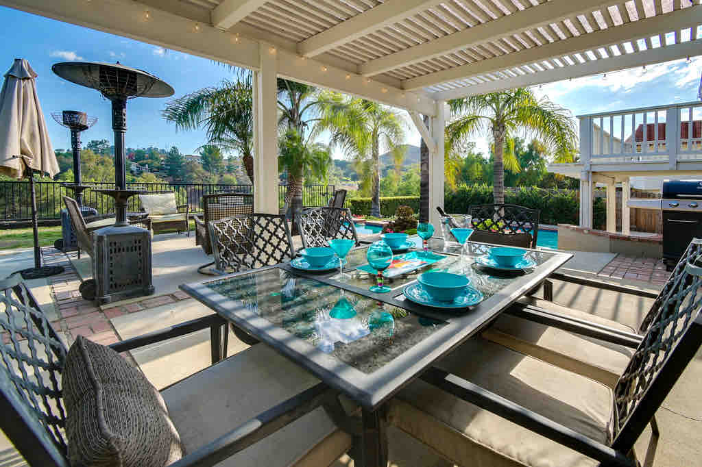 An outdoor dining area features a large table set with turquoise dinnerware, surrounded by comfortable seating. Lush greenery and palm trees provide a vibrant backdrop, while a patio heater stands nearby, creating an inviting atmosphere for al fresco meals.