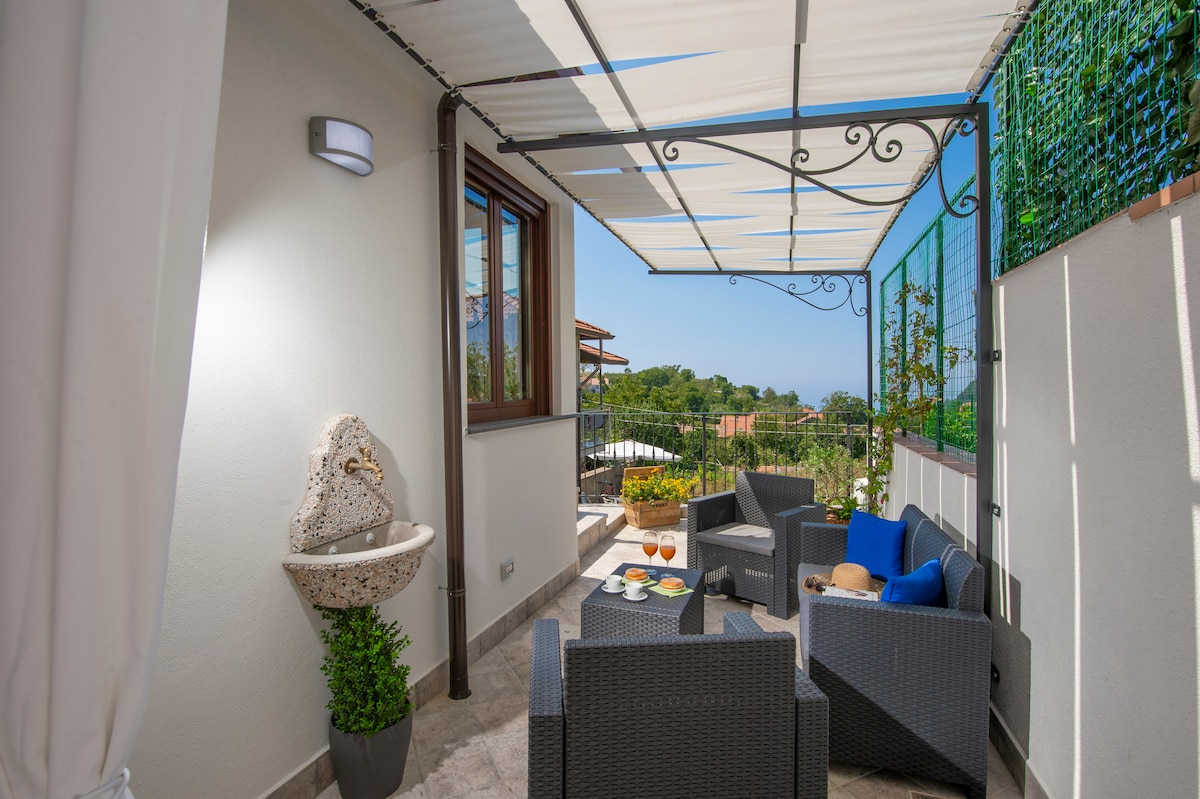 A shaded outdoor seating area is furnished with comfortable chairs and a small table. A decorative water feature and potted plant enhance the ambiance. The surrounding greenery is visible through the window, providing a connection to the natural landscape.