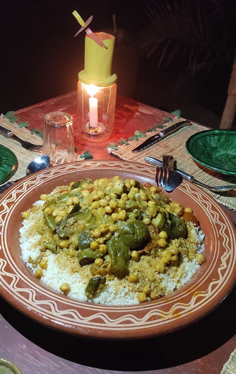 A traditional dish is presented in a large, decorated bowl, featuring a mix of couscous, vegetables, and chickpeas. The table is set with green glass plates and cutlery, illuminated by a candle in a glass holder, creating a cozy dining atmosphere.