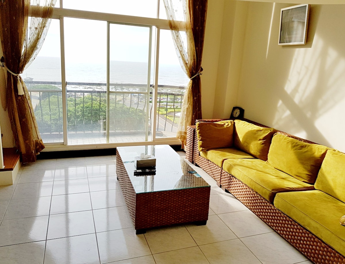 A bright living area features a large window offering ocean views, enhanced by sheer curtains. A green sofa and glass-topped table are arranged on polished tile flooring, creating an open and airy feel.