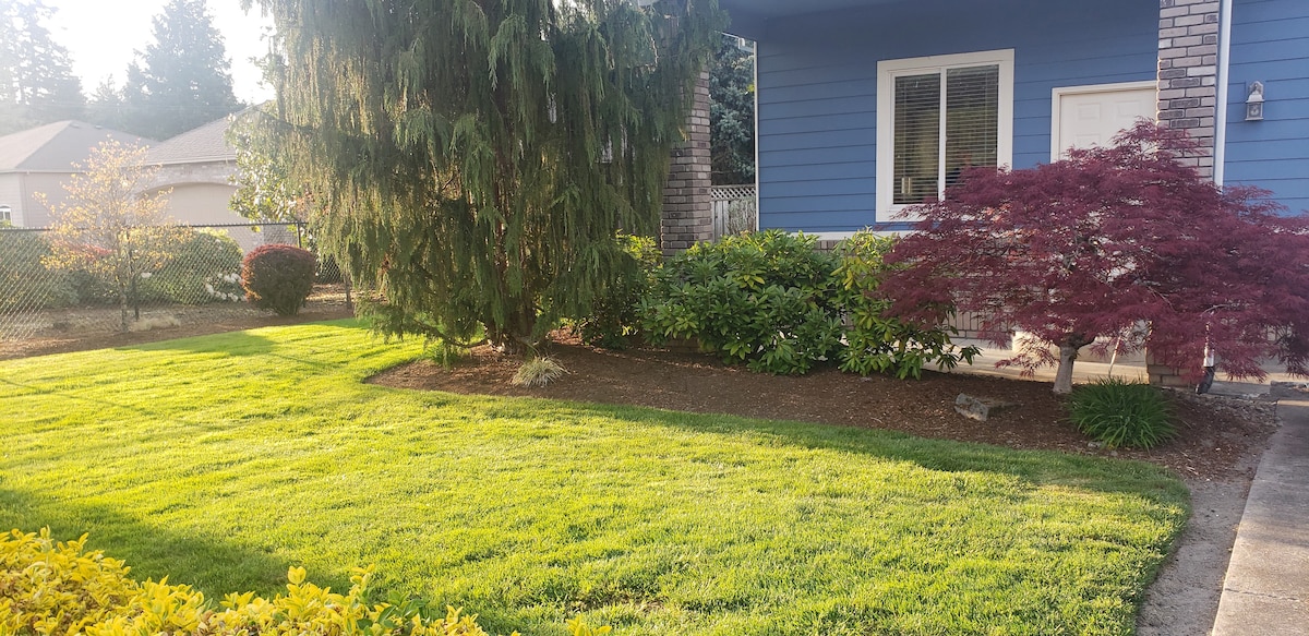 A well-maintained lawn features lush green grass, bordered by neatly trimmed shrubs and small trees. A vibrant red maple adds a pop of color, while the blue exterior of the guest house is visible in the background, framed by the landscaping.