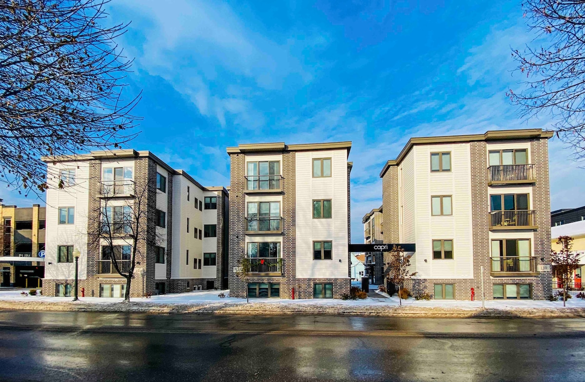 The exterior view of the condo building shows a modern three-story structure with a brick and siding facade. Large windows are present, and some balconies are visible. The surrounding area features light snow, with a clear blue sky overhead.