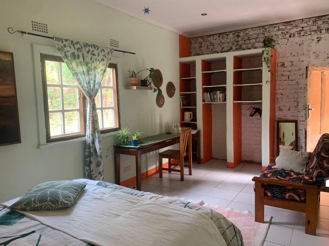 A self-contained room features a spacious bed with decorative linens. A small desk and chair are positioned near a window, with potted plants enhancing the space. Bookshelves and an armchair complete the room's layout, which is accented by a mix of neutral and warm tones.