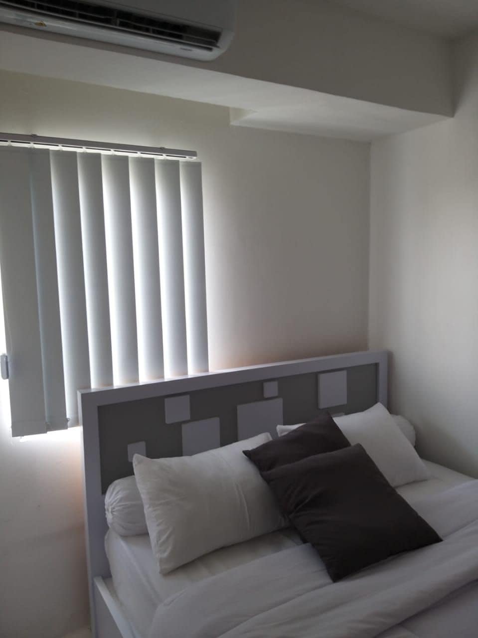 A sleeping area features a spacious bed with several soft pillows arranged on top. Natural light is filtered through vertical blinds covering a nearby window, illuminating the light-colored walls and minimalist headboard.