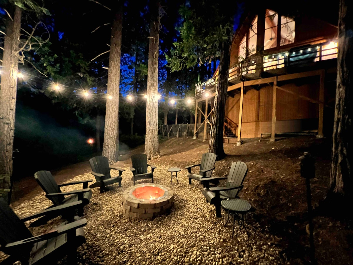 An inviting fire pit area is surrounded by six Adirondack chairs, perfect for evening gatherings. Warm light from string lights casts a cozy glow over the gravel path, while the backdrop of tall trees and the cabin structure create a serene forest setting.