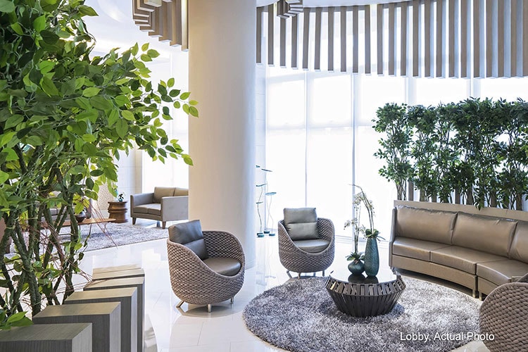 A bright lobby area features large windows allowing ample natural light. Comfortable seating arrangements include modern chairs and a curved sofa, arranged around a decorative round table. Lush green plants provide a refreshing touch, enhancing the inviting ambiance of the space.