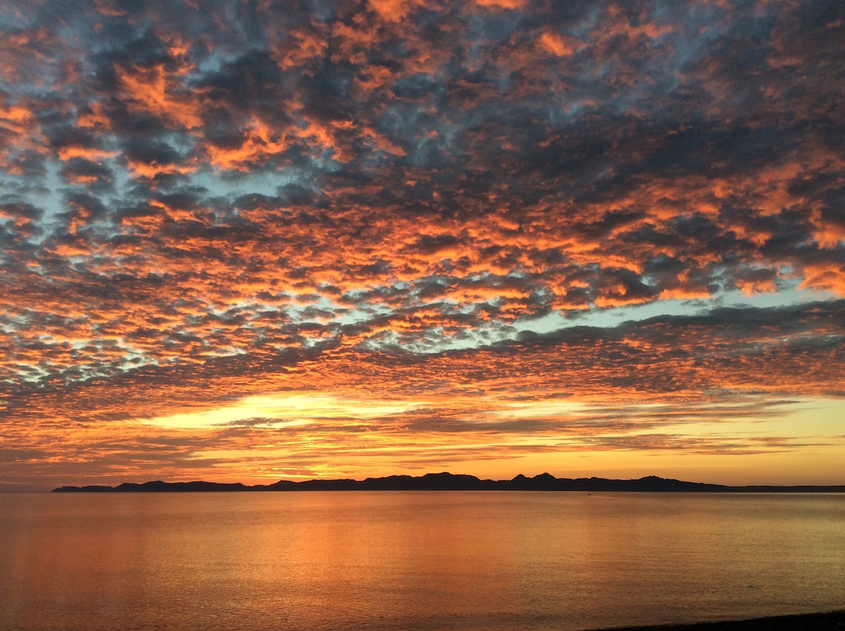 A vibrant sunset paints the sky with shades of orange, pink, and blue, capturing the tranquil Sea of Cortez. The horizon features the silhouette of Carmen Island, while gentle waves create a serene reflection of the colorful sky.