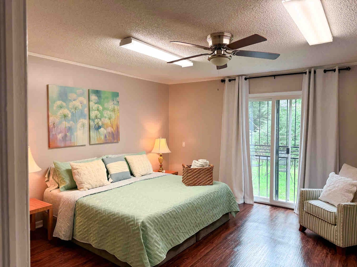 A spacious bedroom features a large bed adorned with soft bedding and two paintings above it. Natural light filters through a set of glass doors that lead to a small balcony. A comfortable chair and nightstands with lamps complete the inviting decor.