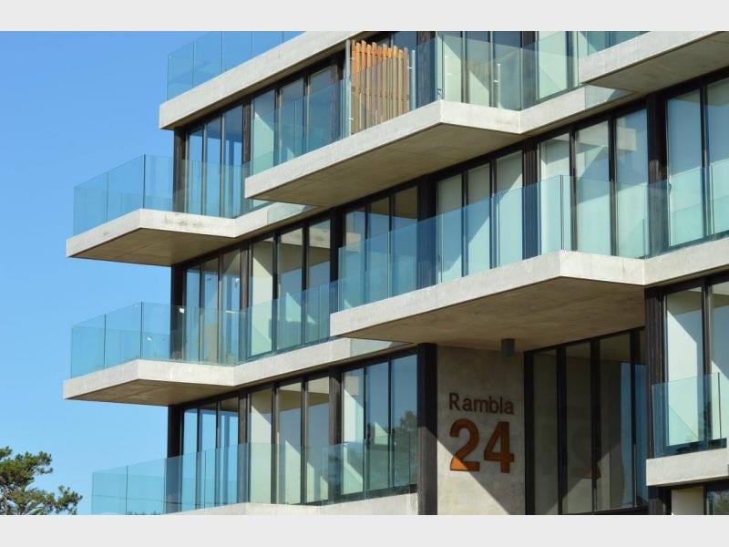 A modern building is showcased with multiple levels, featuring large glass balcony railings that offer unobstructed views. The concrete facade is complemented by the bold address 'Rambla 24', with a clear blue sky in the background enhancing the structure's contemporary design.