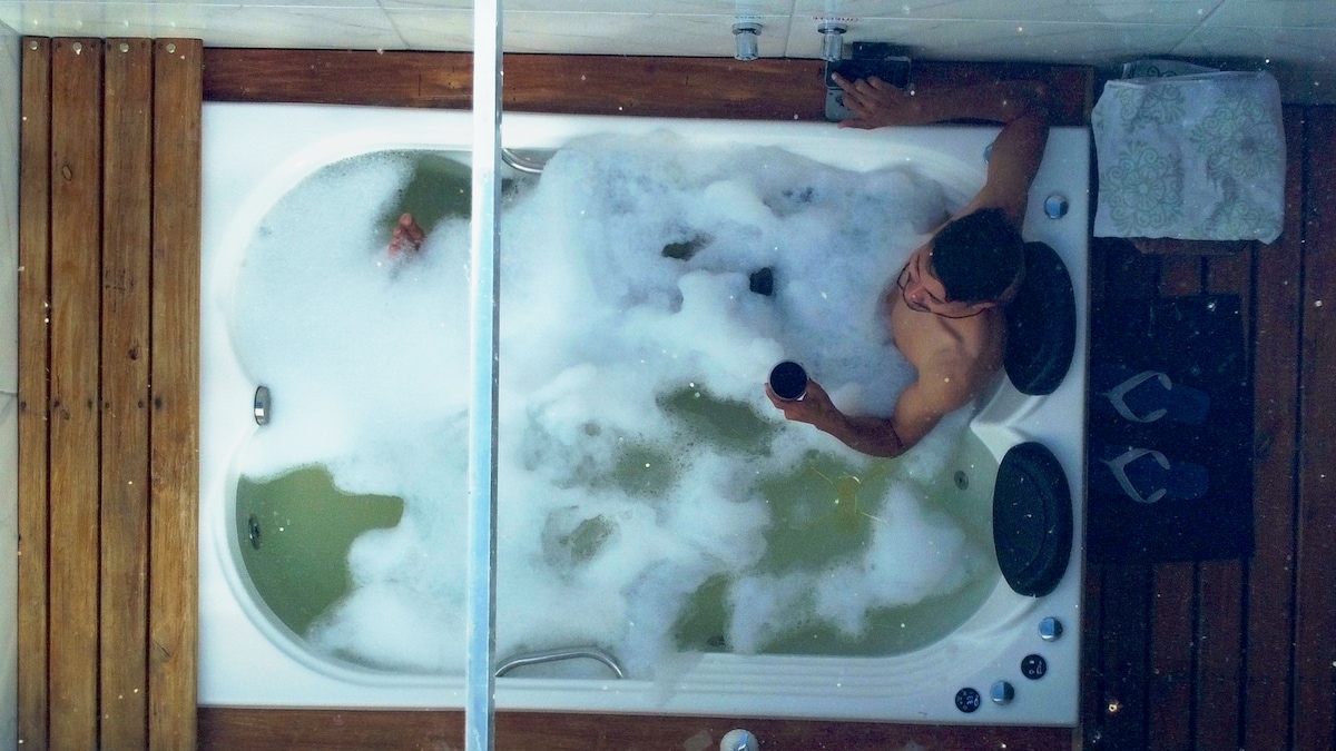 A spacious hydromassage bathtub is filled with bubbles, surrounded by wooden decking. Soft lighting enhances the relaxing atmosphere, while personal items are placed neatly nearby. The overhead view captures a sense of tranquility and invites moments of relaxation.