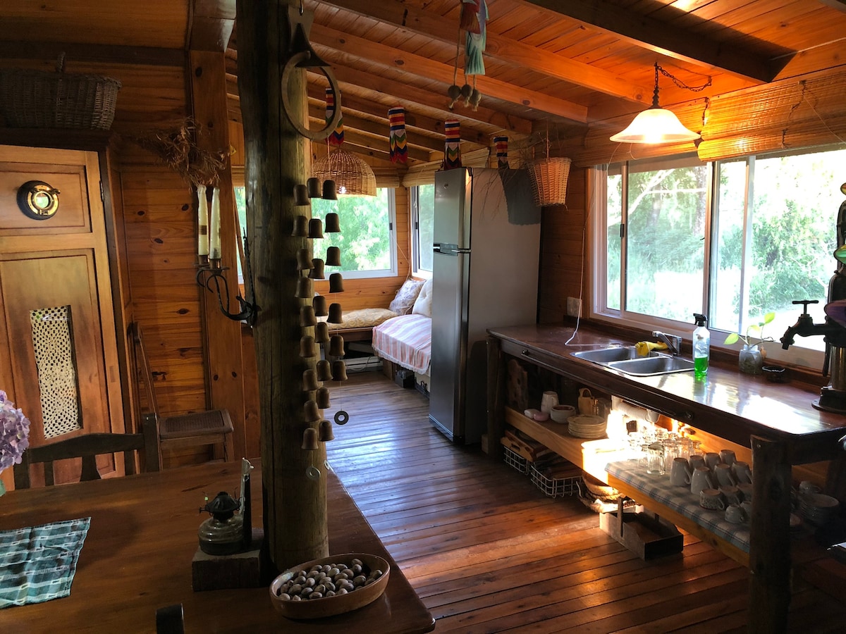 A wooden interior features a cozy kitchen area with a large window allowing natural light. A dining table sits prominently, complemented by decorative elements. A functional refrigerator and an abundance of kitchenware are visible, creating an inviting space for meal preparation.