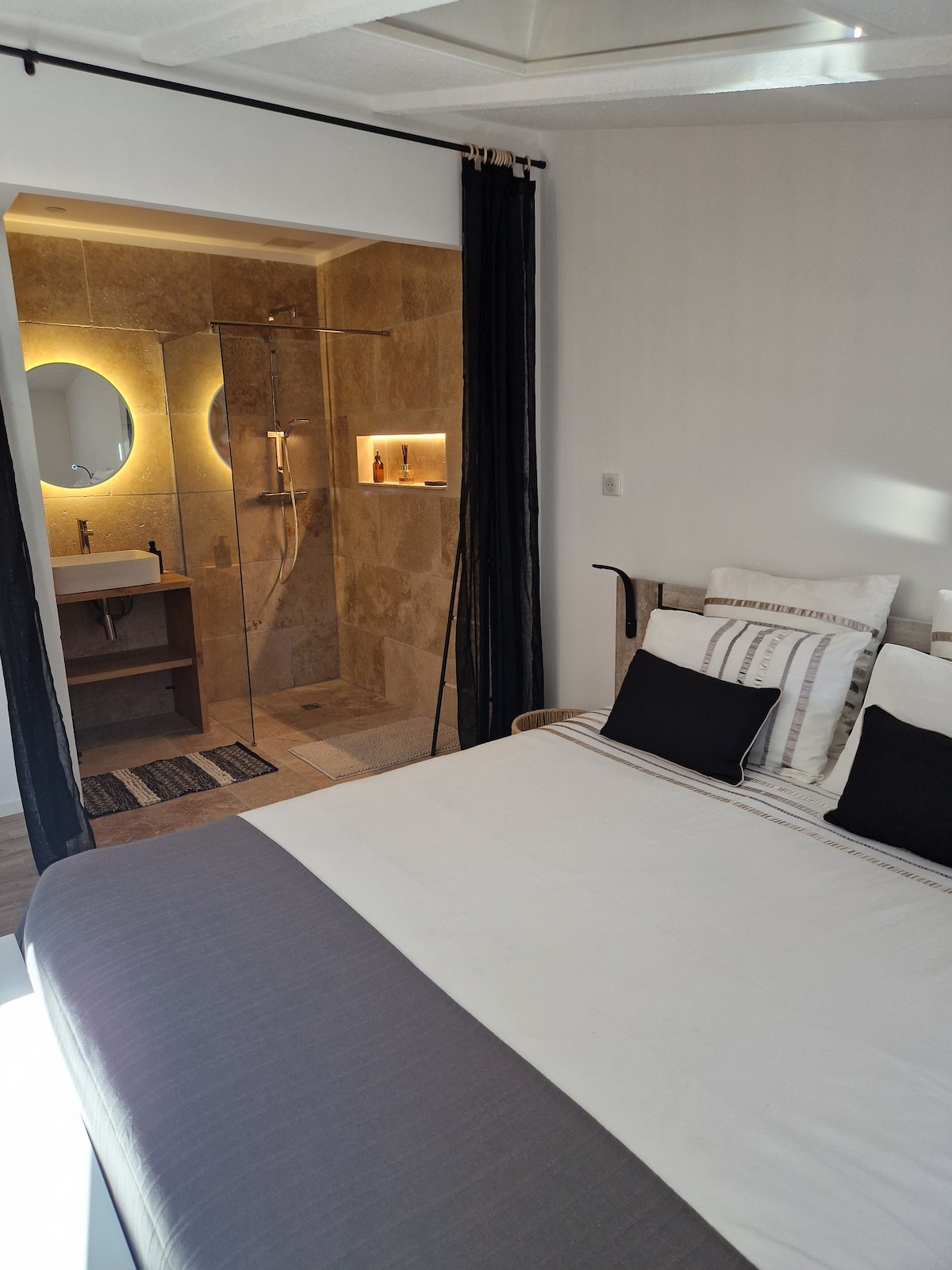 A bright sleeping area features a spacious bed adorned with neutral-toned linens and decorative pillows. The adjoining bathroom is visible, showcasing a glass-enclosed shower with warm lighting and modern fixtures, complemented by stone tiles and a minimalist vanity.