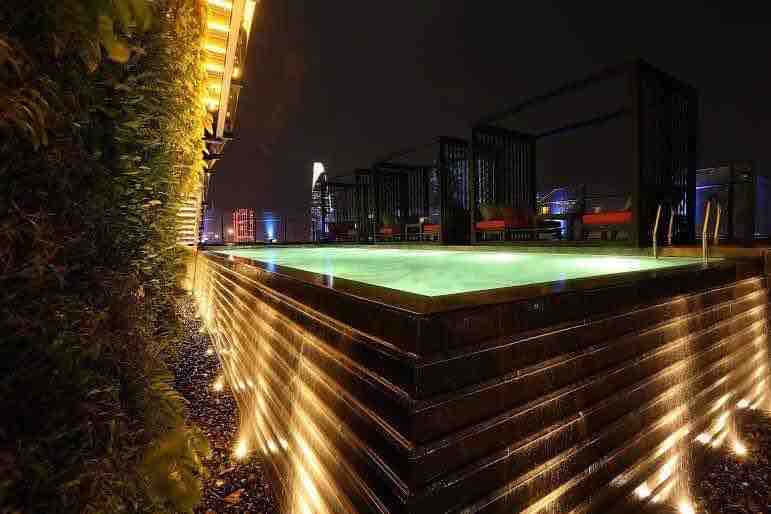 A rooftop pool is illuminated at night, featuring a modern design with cascading water features. Subtle lighting enhances the pool's greenish hue, while lounge areas with black structures are visible in the background, surrounded by city skyline views.