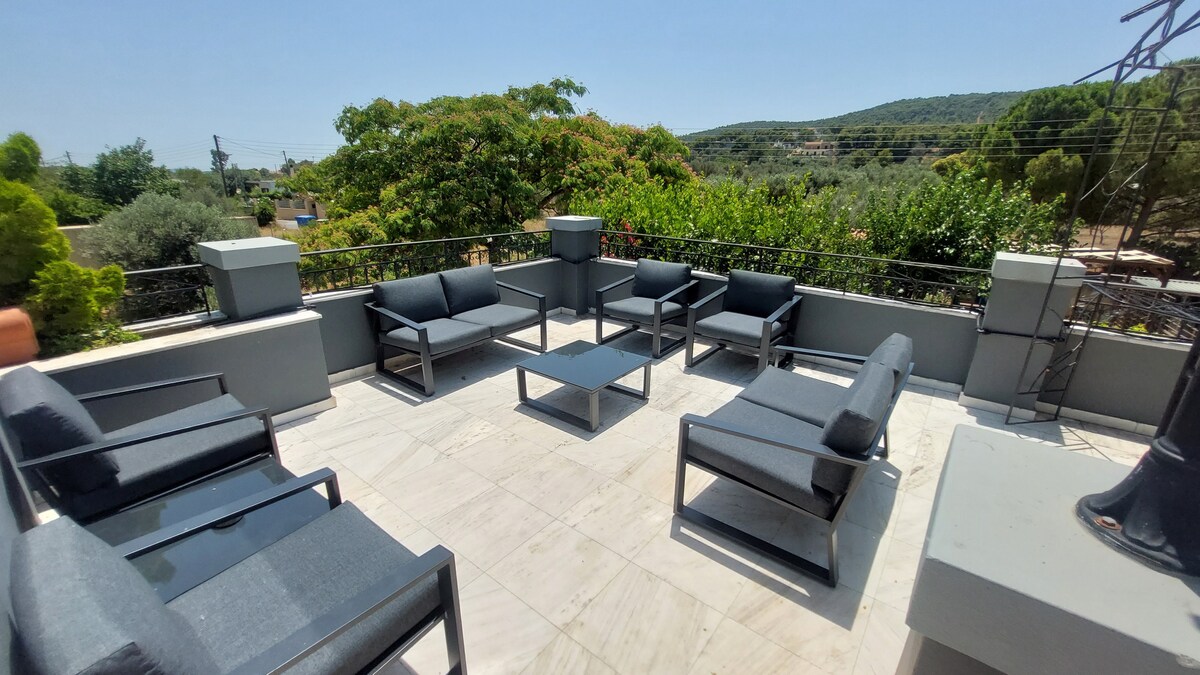 An outdoor terrace features a set of dark lounge furniture arranged around a central coffee table. The area is complemented by views of greenery and distant hills, providing a calm setting for relaxation. Bright sunlight enhances the spacious feel of the terrace.