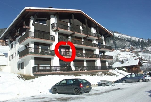 A chalet-style building featuring multiple balconies and wooden accents is surrounded by snow. The structure showcases a welcoming design, and several parked cars are present in front, indicating access to the residence. The serene mountain landscape can be observed in the background.
