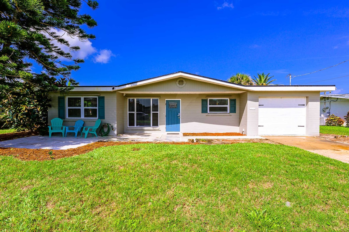 A charming single-story home is presented with a well-manicured lawn, featuring a driveway leading to a garage. Two light blue chairs sit on a concrete patio in front of the house, accented by vibrant greenery and a clear blue sky above.