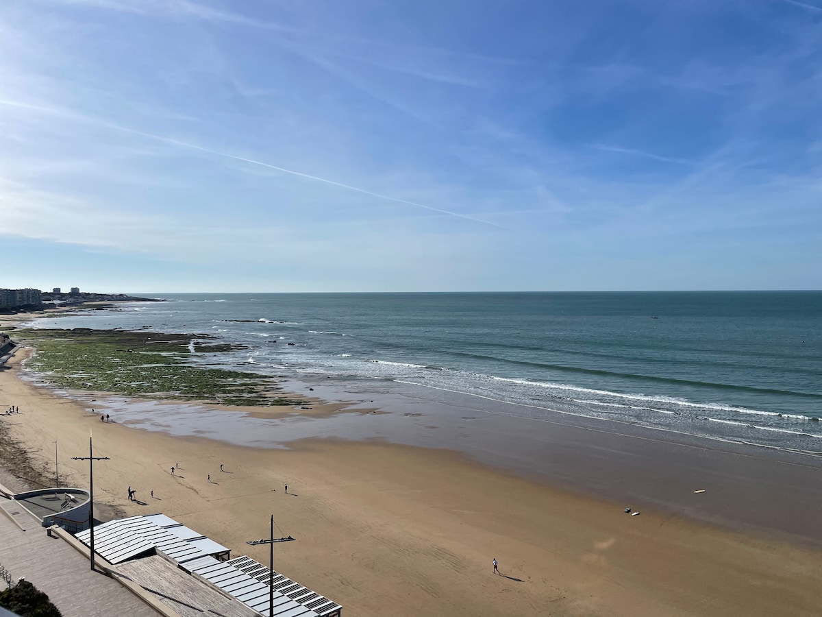 A panoramic view of a spacious beach is displayed, featuring a golden sandy shore and gentle waves lapping against the water. Sunlight reflects on the sea, while distant figures walk along the beach. The sky is clear with light clouds, enhancing the serene atmosphere.