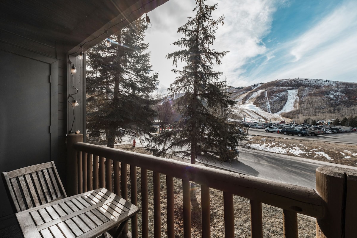 A balcony is shown with wooden furniture facing a scenic view of the surrounding mountains and ski slopes. Tall pine trees frame the image, while a road and parking area are visible in the foreground, hinting at accessibility to outdoor activities.