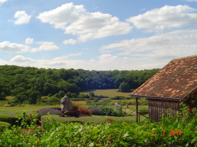 A serene landscape is captured, featuring rolling green hills bordered by dense forests. In the foreground, well-maintained hedges are visible, complemented by flowering plants. A rustic wooden building with a tiled roof is positioned to the right, adding to the peaceful rural scenery.