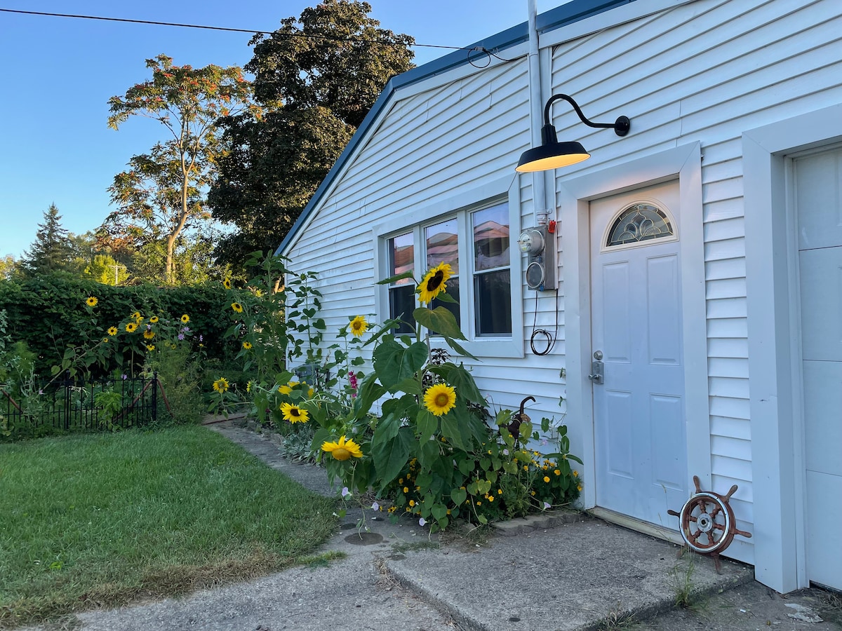 The exterior of the cottage features a white panel facade along with a welcoming door, surrounded by a garden of vibrant sunflowers and lush greenery. A light fixture hangs above the entrance, illuminating the pathway leading to the door.