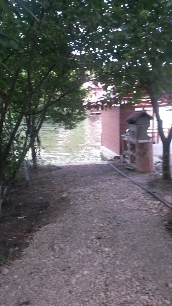 A pathway bordered by trees leads to the water's edge, indicating proximity to the river. A wooden structure is partially visible, suggesting a dock or pier for water activities. The area is tranquil and natural, creating a serene atmosphere.