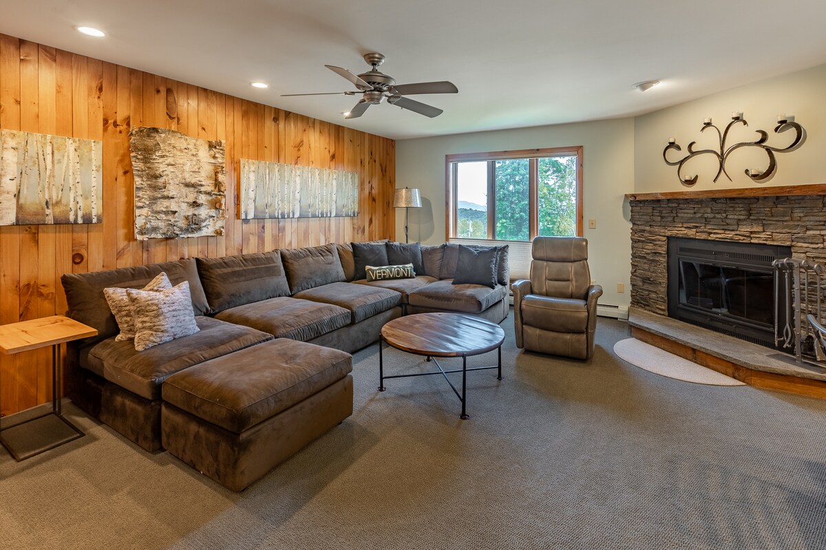 A cozy living area features a large sectional sofa against a wood-paneled wall, complemented by decorative art pieces. A stone fireplace is situated beneath an elegantly designed mantel, with a large window allowing natural light to brighten the space. A small side table and a recliner enhance the seating options.