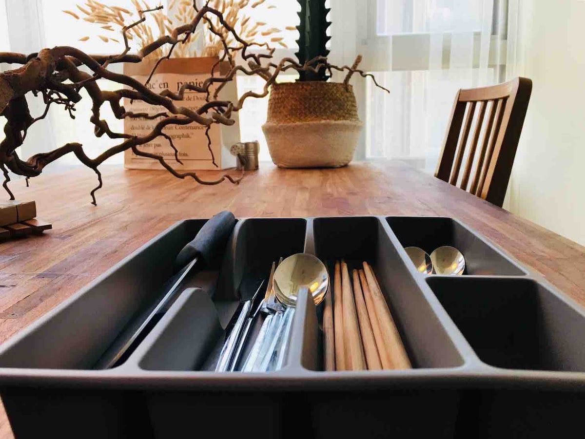 A neatly arranged utensil organizer sits on a wooden table, showcasing various silverware and chopsticks. The background features a decorative arrangement of branches in a pot, accompanied by a woven basket, adding texture to the dining space.