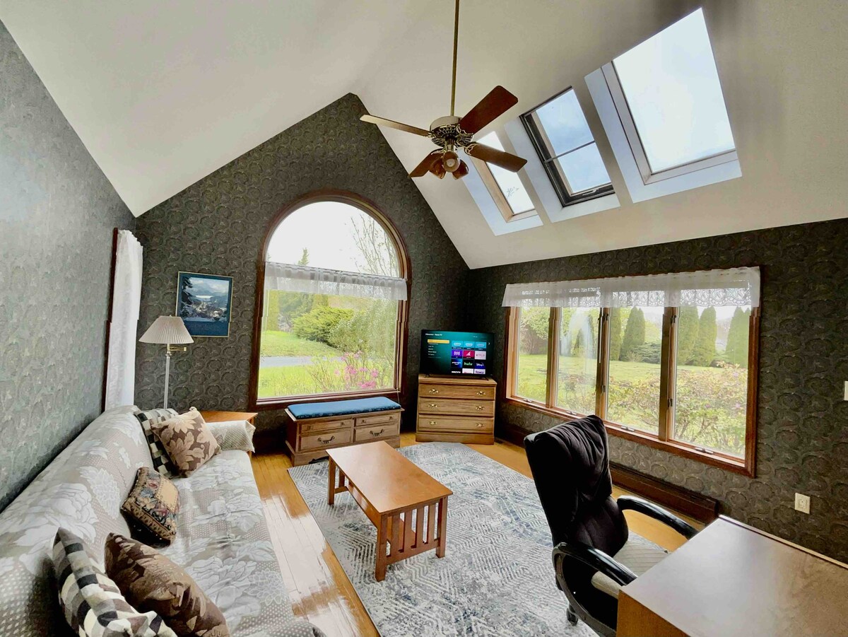 A spacious living area is highlighted by large windows and skylights that allow natural light to fill the room. A comfortable sofa is positioned alongside a wooden coffee table, while a television and a wooden dresser add functionality. Outdoor greenery is visible through the windows.
