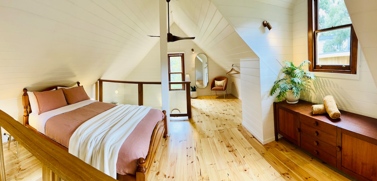 The airy loft area features a spacious bed with soft pink bedding, complemented by natural wood floors and light walls. A ceiling fan hangs above, and windows offer natural light. A small plant sits atop a dark wood dresser, adding a touch of greenery.