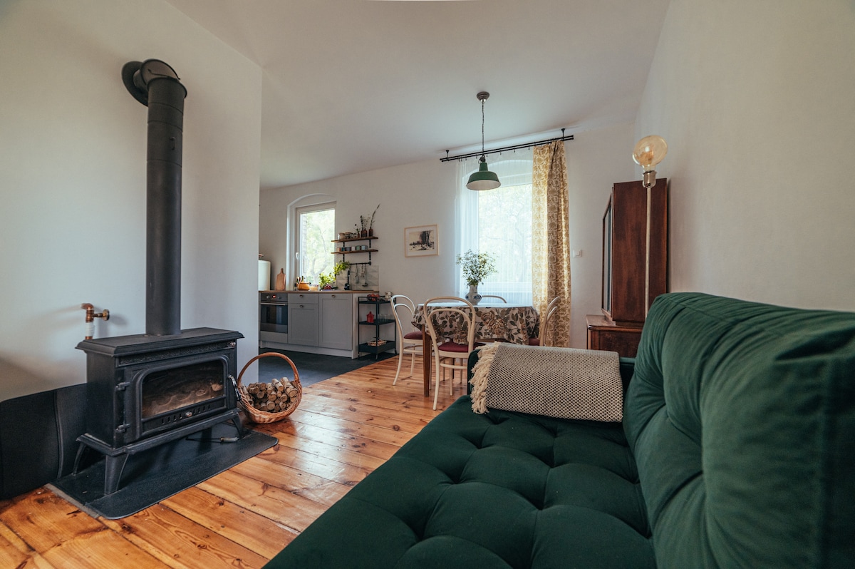 The interior features a cozy living area with a green sofa positioned near a black wood stove. A dining table with white chairs and a decorative floral area can be seen in the background, complemented by a bright window and warm wood flooring.